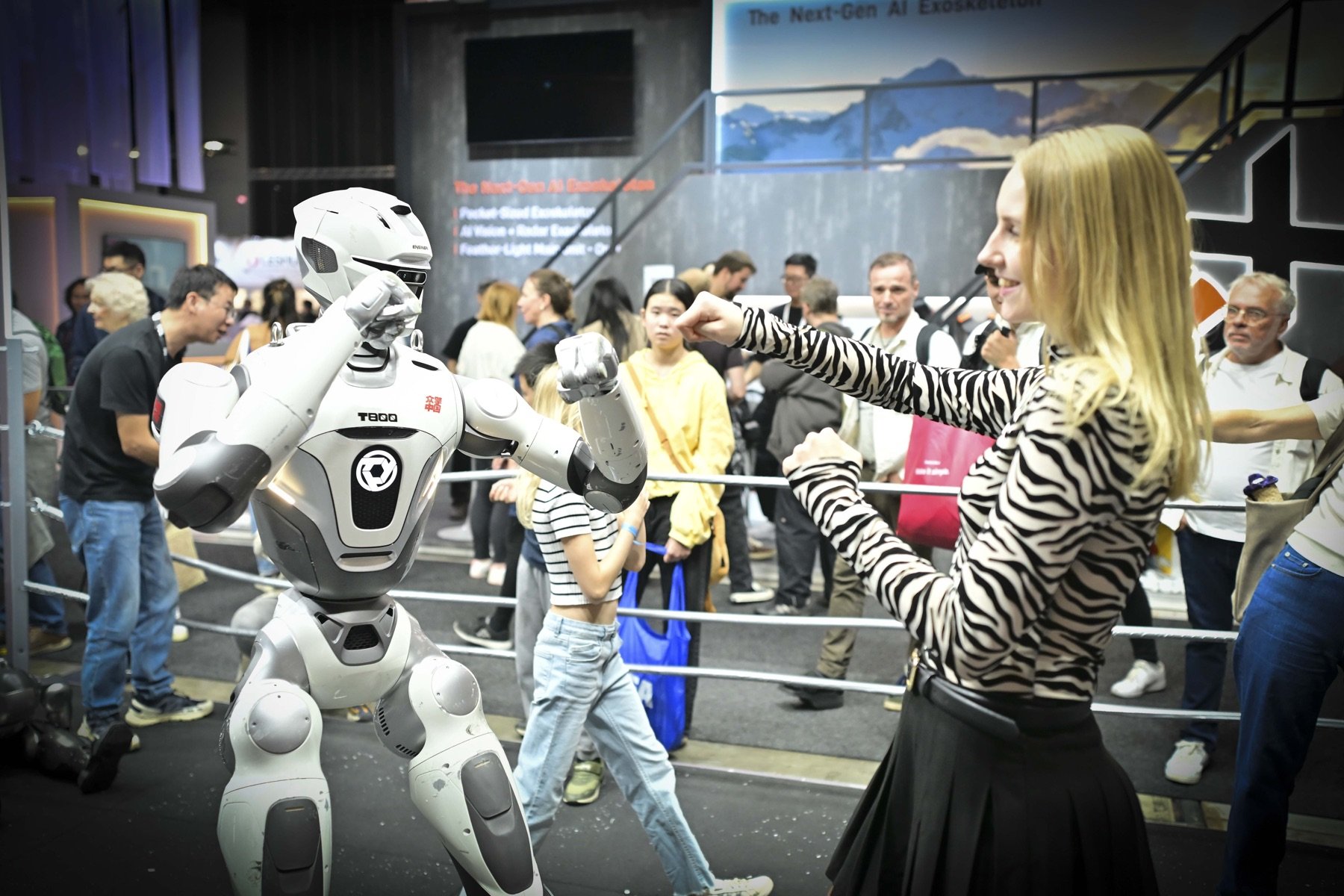 Ein Roboter boxt im Ring gegen eine Besucherin, dahinter Werbung für ein KI-Exoskelett